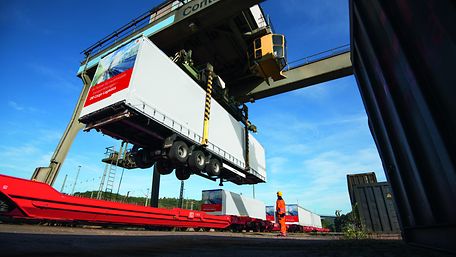 A semi-trailer is loaded onto a train by a gantry crane at a transhipment terminal